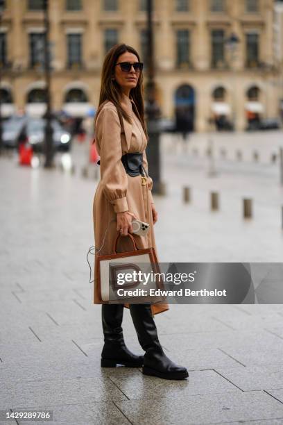 Guest wears black sunglasses, gold earrings, a brown long coat, a black shiny leather Saddle large belt from Dior, a white latte fabric and brown...