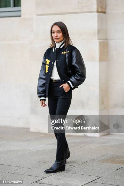 Guest wears a white turtleneck wool pullover, a black felt and leather sleeves with embroidered yellow yoke pattern bomber coat, a black shiny...