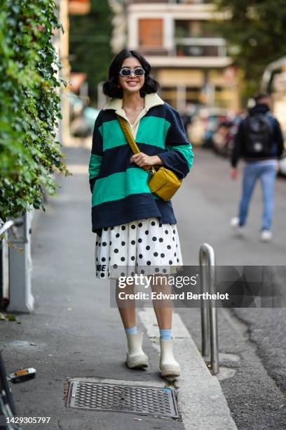 Guest wears blue sunglasses, a pale yellow collar / navy blue and green large striped print patten oversized polo shirt, a yellow nylon crossbody...