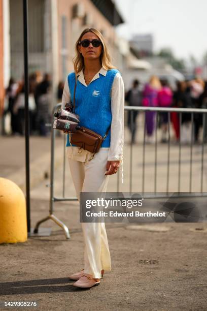 Guest wears black sunglasses from Ray Ban, earrings, a gold necklace, a pale yellow shirt, a blue braided wool sleeveless with white embroidered logo...