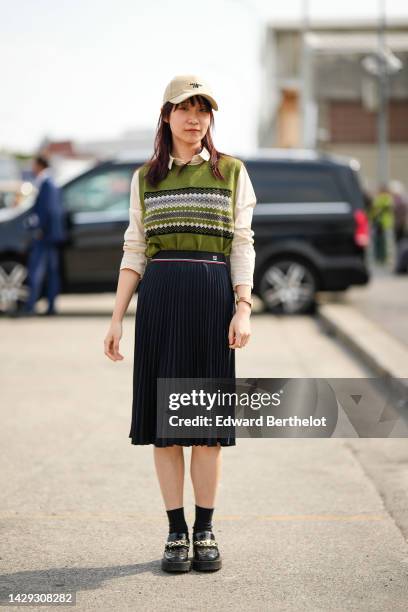 Guest wears a beige denim with black embroidered logo cap, a beige shirt, a green with black and white print pattern sleeveless wool pullover, a...