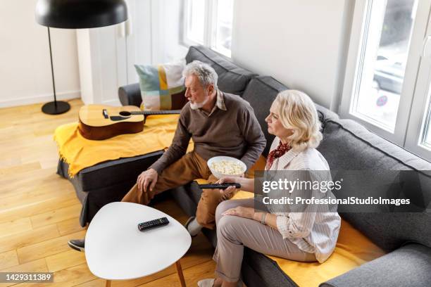 an older couple is sitting on a sofa at home, watching a television program and enjoying popcorn. - city of joy premiere stock pictures, royalty-free photos & images