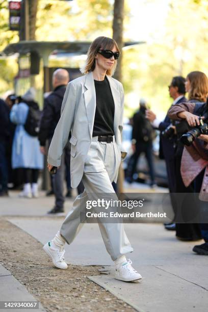 Anya Ziourova wears black sunglasses, a black t-shirt, a pale gray blazer jacket, matching pale gray large suit pants, white latte socks, white shiny...