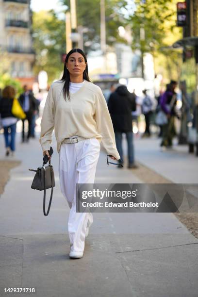 Guest wears a white t-shirt, a beige wool pullover, a white latte leather belt, white suit pants, a black shiny leather Kelly handbag from Hermes,...