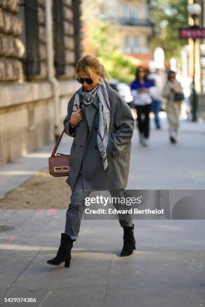 Guest wears black sunglasses, a white and black print pattern silk scarf, a gray oversized jacket, gray denim pants, a brown shiny leather handbag,...