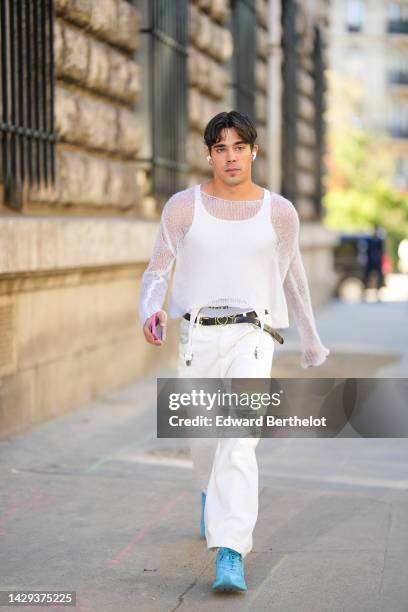 Guest wears gold earrings, a white wool transparent pullover, a white tank-top, a black shiny leather with gold buckle belt, white denim large pants,...