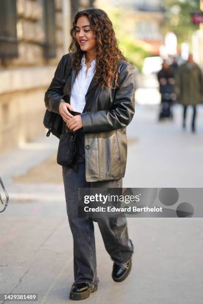 Guest wears a white shirt, a black shiny leather buttoned blazer jacket, a black shiny leather shoulder bag, black denim large pants, black shiny...