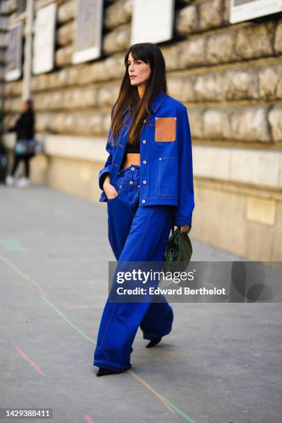 Leia Sfez wears a black cropped top, a royal blue denim with brown leather embossed pattern shirt from Loewe, matching royal blue denim large pants...