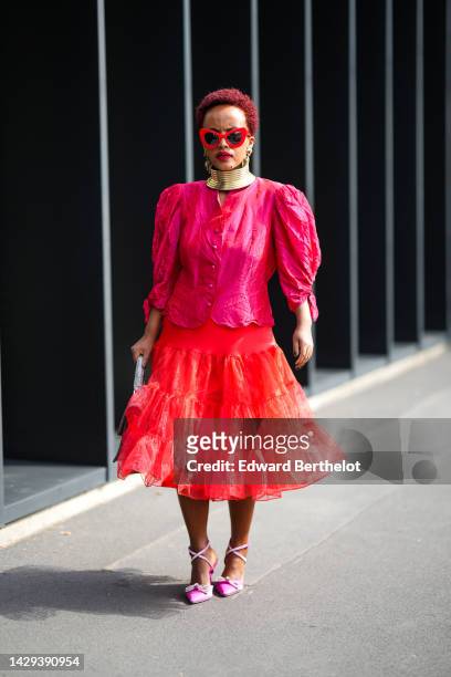 Guest wears red sunglasses, a gold large necklace, a neon pink puffy sleeves / buttoned shirt, a neon red shiny tulle ruffled midi skirt, a grey...