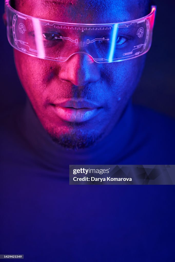 A young African-American man in futuristic fashion glasses with neon light looks at camera. The concept of the future, computer games, augmented reality, virtual reality, modern technologies, technological progress, escapism.