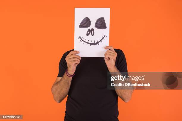 man covering his face with a paper in which a smiling and terrifying halloween face is painted, on an orange background. concept of celebration, day of the dead and carnival. - covering nose stock pictures, royalty-free photos & images