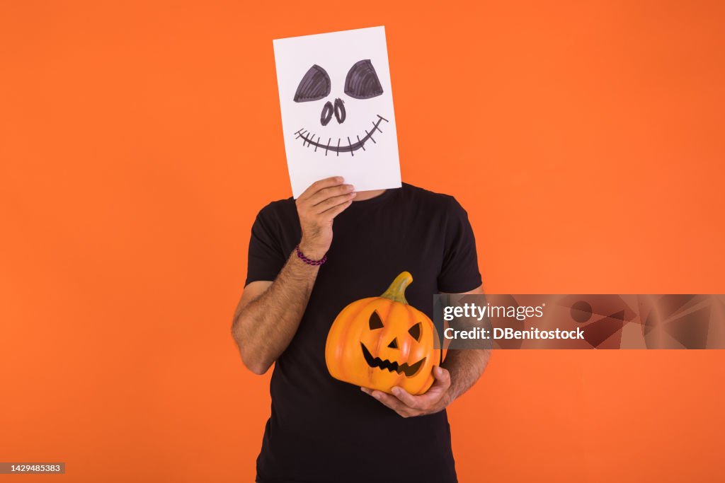 Man covering his face with a paper on which a terrifying smiling Halloween face is painted, holding a pumpkin, on orange background. Concept of celebration, day of the dead and carnival.