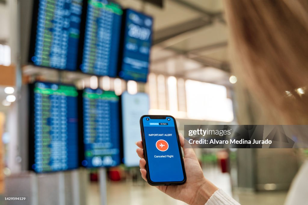 Close-up view of an unrecognizable woman's hand in airport terminal holding her mobile phone receiving a notification with her flight cancellation.