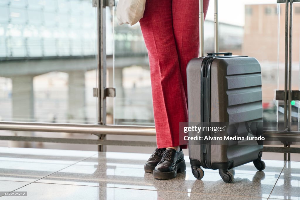 Detail view of unrecognizable woman's legs next to a suitcase at the airport terminal waiting for a flight departure.