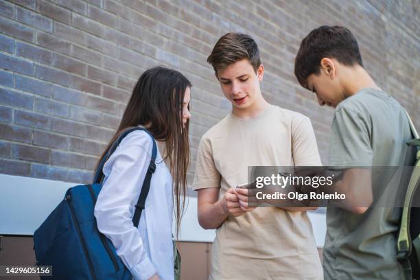 cyberbullying by messages and hypocrisy in high school. students surfing mobile internet, social net near school - 3 teenagers mobile outdoors stockfoto's en -beelden