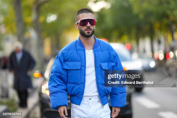 Guest wears red futurist sunglasses, a white t-shirt, a gold long chain pendant necklace, a blue puffer jacket, white denim pants, outside Acne,...