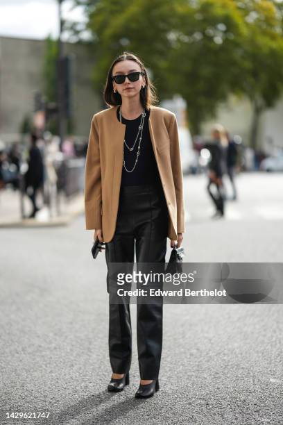 Beatrice Gutu wears black sunglasses, silver earrings, a silver large chains necklace from Hermes, a black t-shirt, a brown felt buttoned blazer...