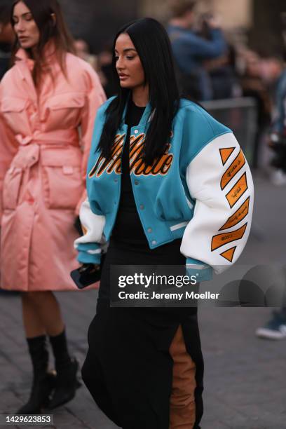 Amina Muaddi is seen wearing a black long slit dress, a blue/white/orange Off-White oversize college bomber jacket and brown suede leather high...