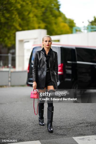 Caroline Daur wears a black ruffled V-neck shirt, a black shiny leather blazer jacket, a black shiny leather large belt, black shorts, black high...