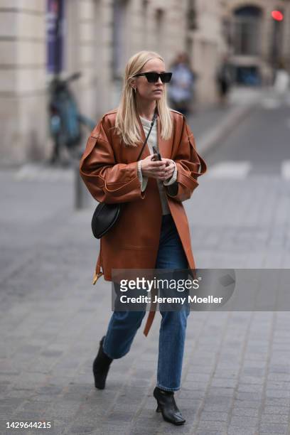 Guest is seen wearing black Saint Laurent sunglasses, gold earrings, creme white wool knit pullover, brown cognac leather blazer jacket, blue denim...