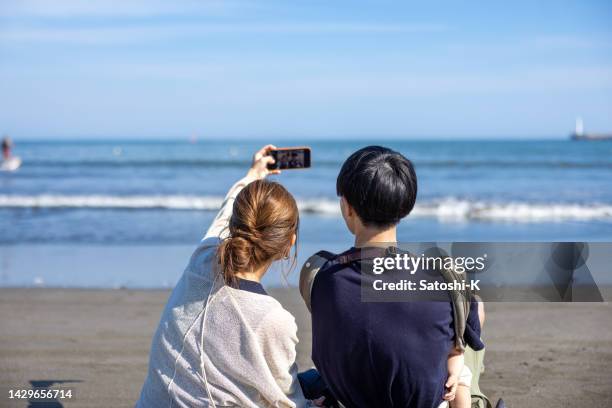 rear view of family sitting on beach, taking selfie pictures - japanese couple beach stock pictures, royalty-free photos & images