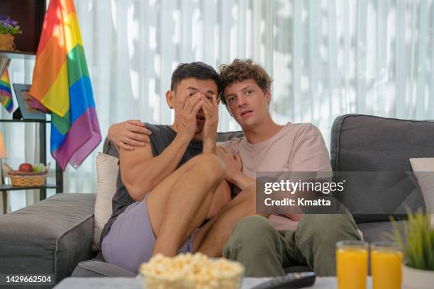 gay men couple watching horror movie on sofa at home, eating popcorn. - horror foto e immagini stock