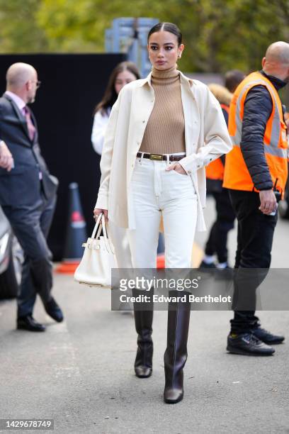 Guest wears gold earrings, a brown ribbed wool turtleneck pullover, a white wool long coat, a dark brown shiny leather belt, white skinny denim...