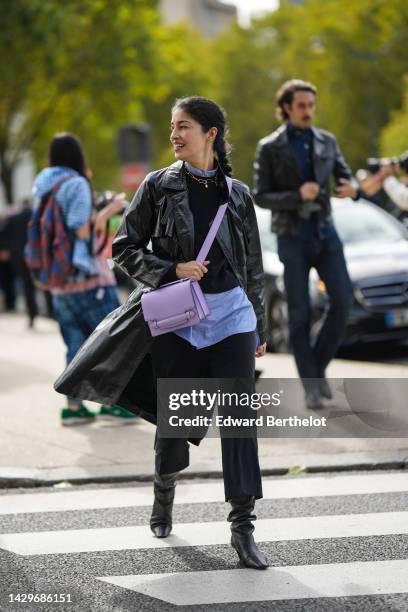 Guest wears silver earrings, a pale blue shirt, a gold large chain necklace, a black wool pullover, a black shiny varnished leather long coat, black...