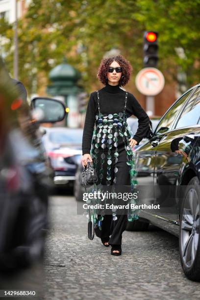 Guest wears black sunglasses, a black turtleneck pullover, a green and white large sequined square-neck / long slit dress, black flared pants, a...