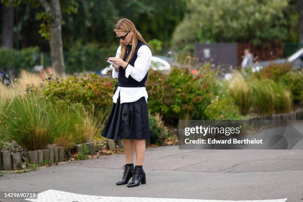 Guest wears black sunglasses, a gold long chain necklace, a navy blue wool sleeveless pullover, rings, a black shiny leather shoulder bag, a black...