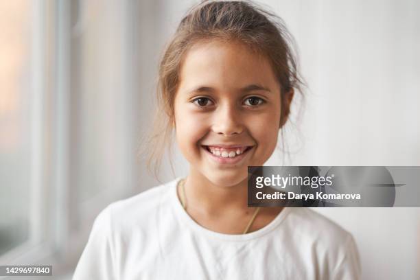 a beautiful brunette girl in a white t-shirt is standing, smiling and looking straight into the camera. a happy child of 8 years old. the concept of a happy childhood. - 8 9 years stock-fotos und bilder