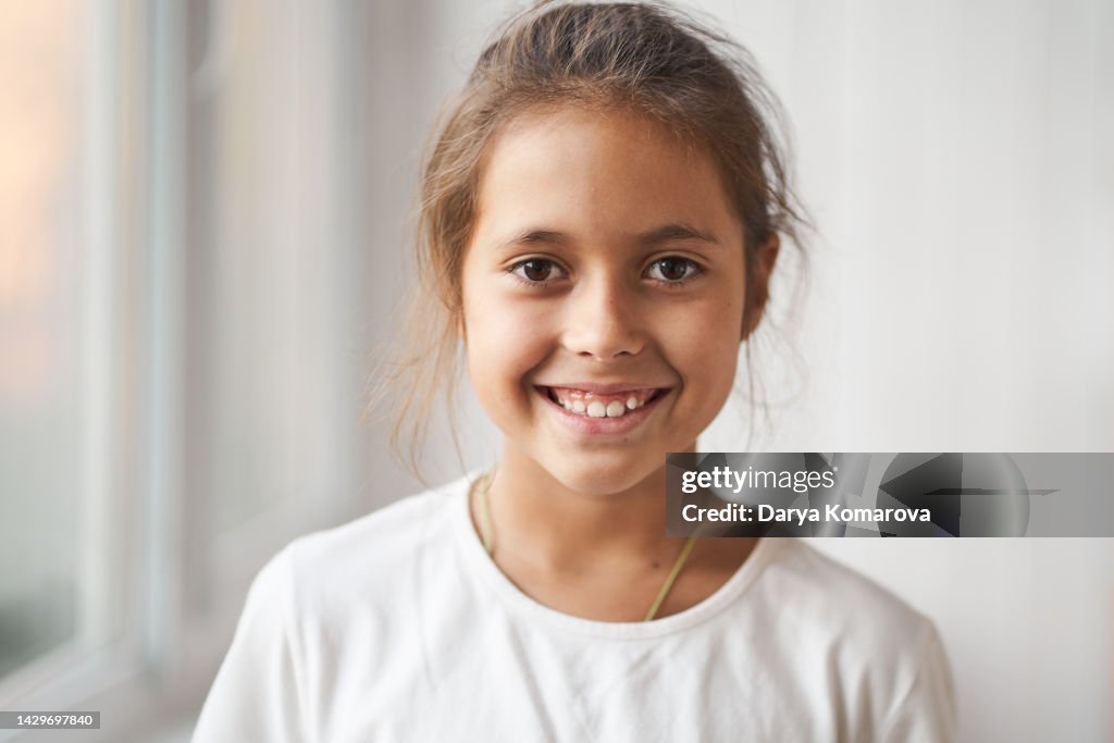 A beautiful brunette girl in a white T-shirt is standing, smiling and looking straight into the camera. A happy child of 8 years old. The concept of a happy childhood.