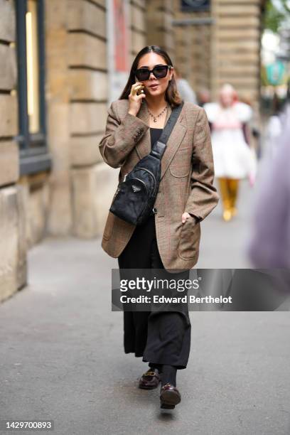 Guest wears black sunglasses, a gold large chain pendant necklace, a black top, a brown checkered print pattern long wool blazer jacket, a black...