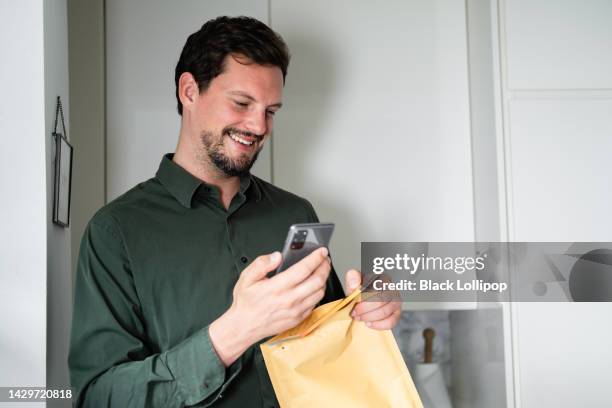 un homme ouvrant une enveloppe, tenant son téléphone intelligent, souriant. - enveloppe matelassée photos et images de collection