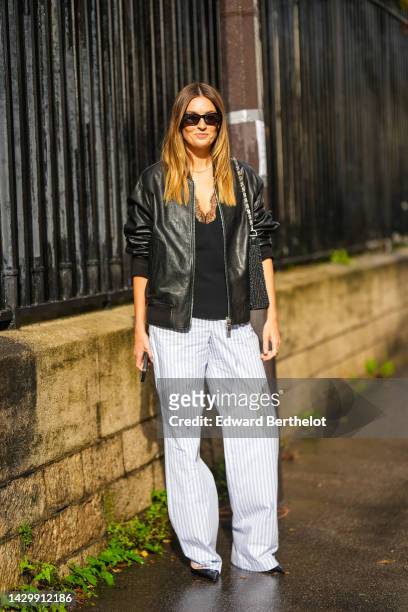 Camille Charriere wears black sunglasses, a white pearls necklace, a black V-neck / lace print pattern tank-top, a black shiny leather jacket, white...