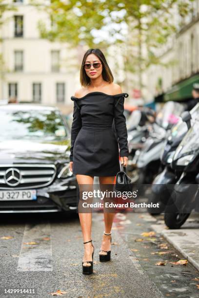 Aimee Song wears black sunglasses, a black shoulder-off / long sleeves short dress, a black shiny leather handbag from Valentino, black shiny...