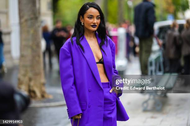 Amina Muaddi wears gold and diamonds earrings, a neon purple oversized blazer jacket, neon purple large wide legs pants, a black bra underwear, a...