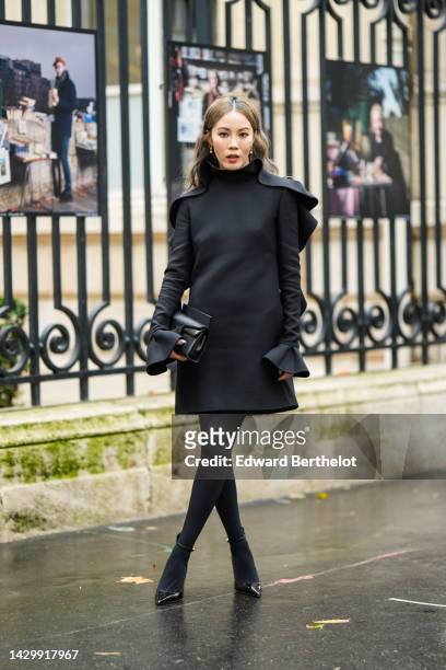 Jenny Tsang in Suet wears gold earrings, a black wavy high neck / long sleeves short dress, a black shiny leather clutch from Valentino, black...