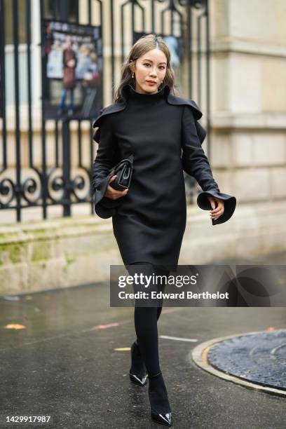 Jenny Tsang in Suet wears gold earrings, a black wavy high neck / long sleeves short dress, a black shiny leather clutch from Valentino, black...