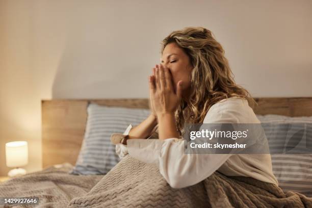 young woman sneezing on a bed in bedroom. - cold virus stock pictures, royalty-free photos & images