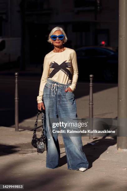 Grece Ghanem wearing blue baggy jeans tight with a white rope, light beige long sleeve top with black hands drawn on it, white shoes, black bag and...