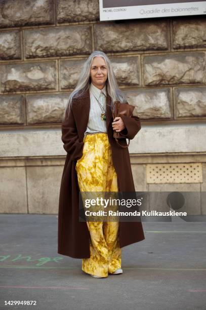 Guest with long grey hair wearing a grey shirt, yellow wide pants with a flower pattern, a long brown coat, and a brown bag outside Loewe during...