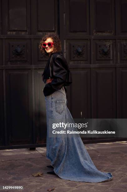 Renia Jaz wearing a black leather cropped jacket, a white and beige body print pattern top, a blue denim long ground touching maxi skirt, a blue...