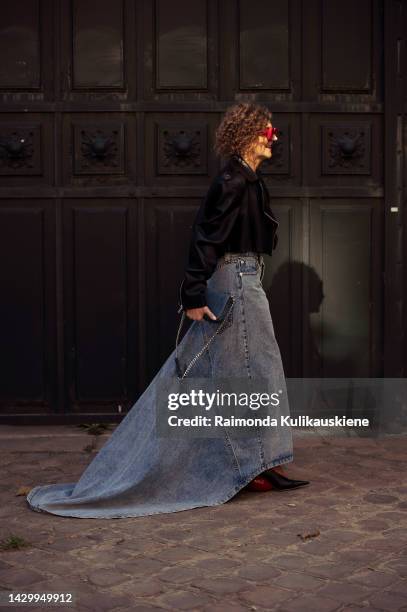 Renia Jaz wearing a black leather cropped jacket, a white and beige body print pattern top, a blue denim long ground touching maxi skirt, a blue...