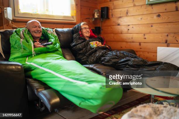 pareja de ancianos tratando de ahorrar costos de energía viendo televisión covoered en sacos de dormir calientes - saco de dormir fotografías e imágenes de stock