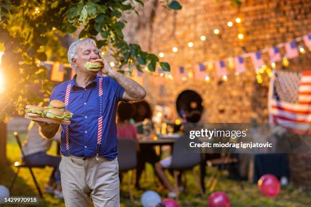 senior man eating a hamburger on celebration of american holiday barbecue party - women in suspenders 個照片及圖片檔