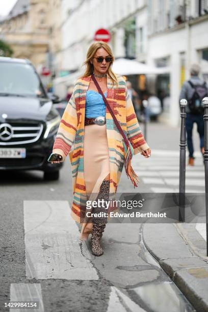 Guest wears brown sunglasses, a silver and blue stones necklace, a brown braided macrame fringed crossbody bag, a yellow / brown / green print...