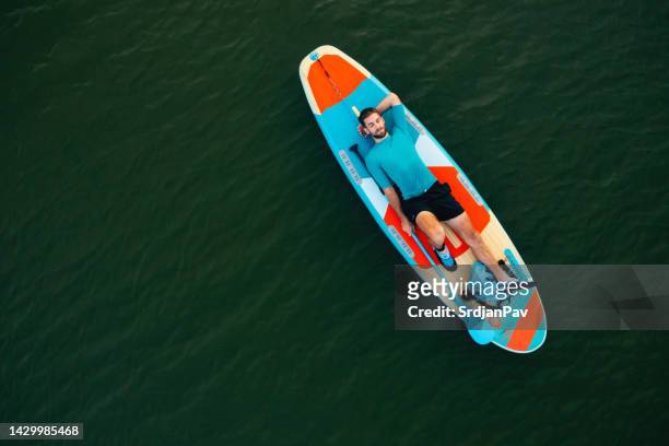 drohnen-gesichtsansicht eines jungen mannes, der auf einem standup-paddleboard liegt - paddleboard stock-fotos und bilder