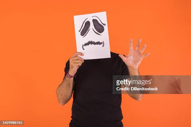 man covered his face with a paper on which a scared halloween face is painted, scaring with his hand, on an orange background. concept of celebration, day of the dead and carnival. - cover monster face stock pictures, royalty-free photos & images