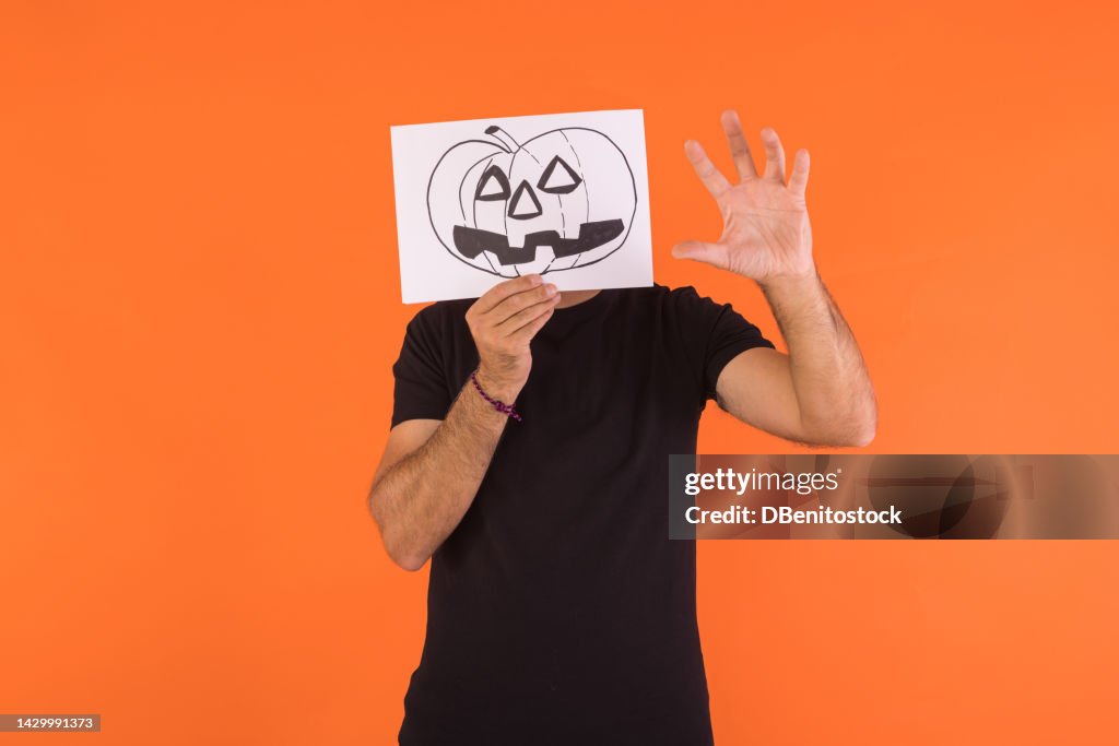 Man covering his face with a paper on which a Halloween Jack o lantern pumpkin is painted, scaring with his hand, on an orange background. Concept of celebration, day of the dead and carnival.
